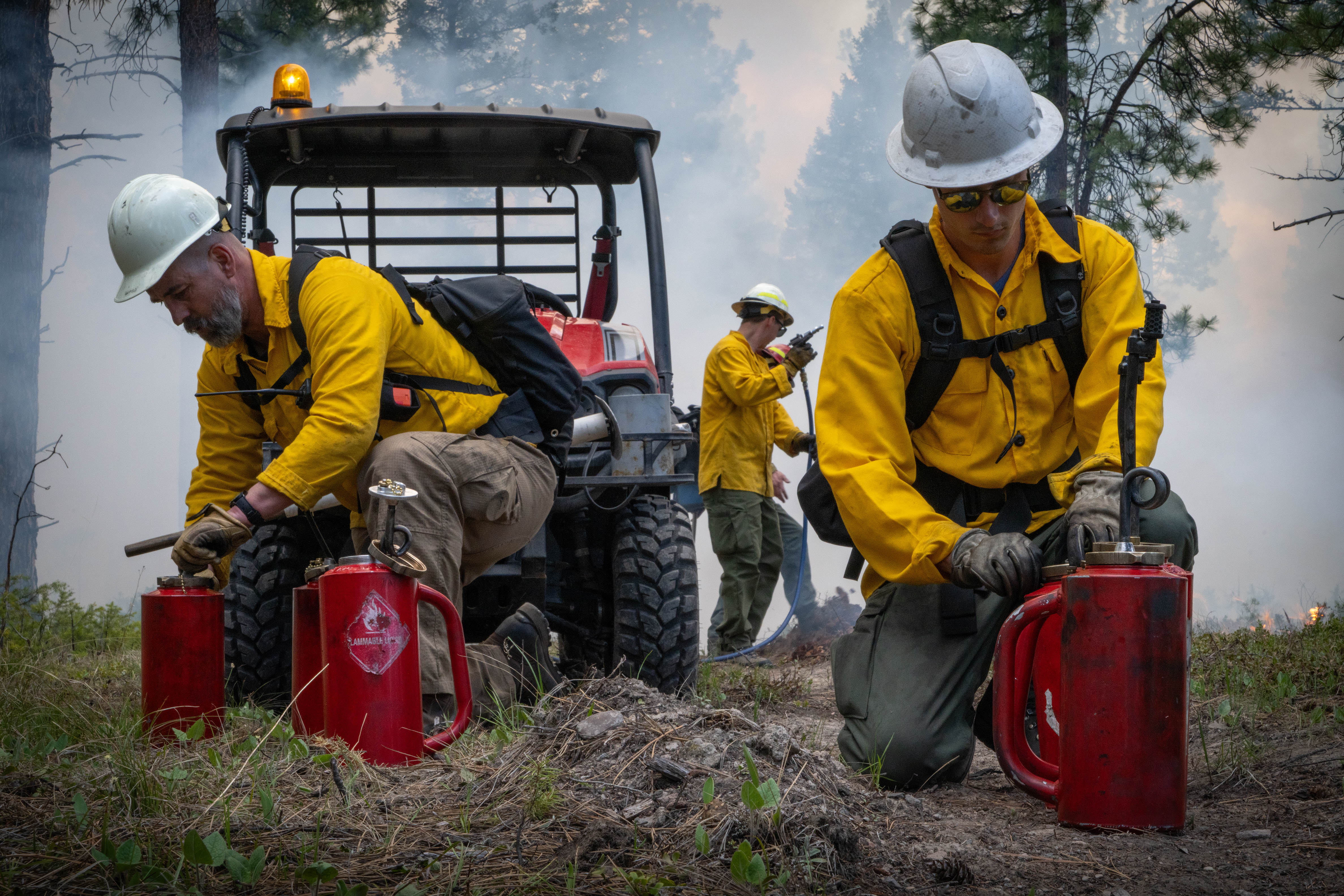 A firefighter wearing full gear checks a drip torch with smokey forest scene in background.