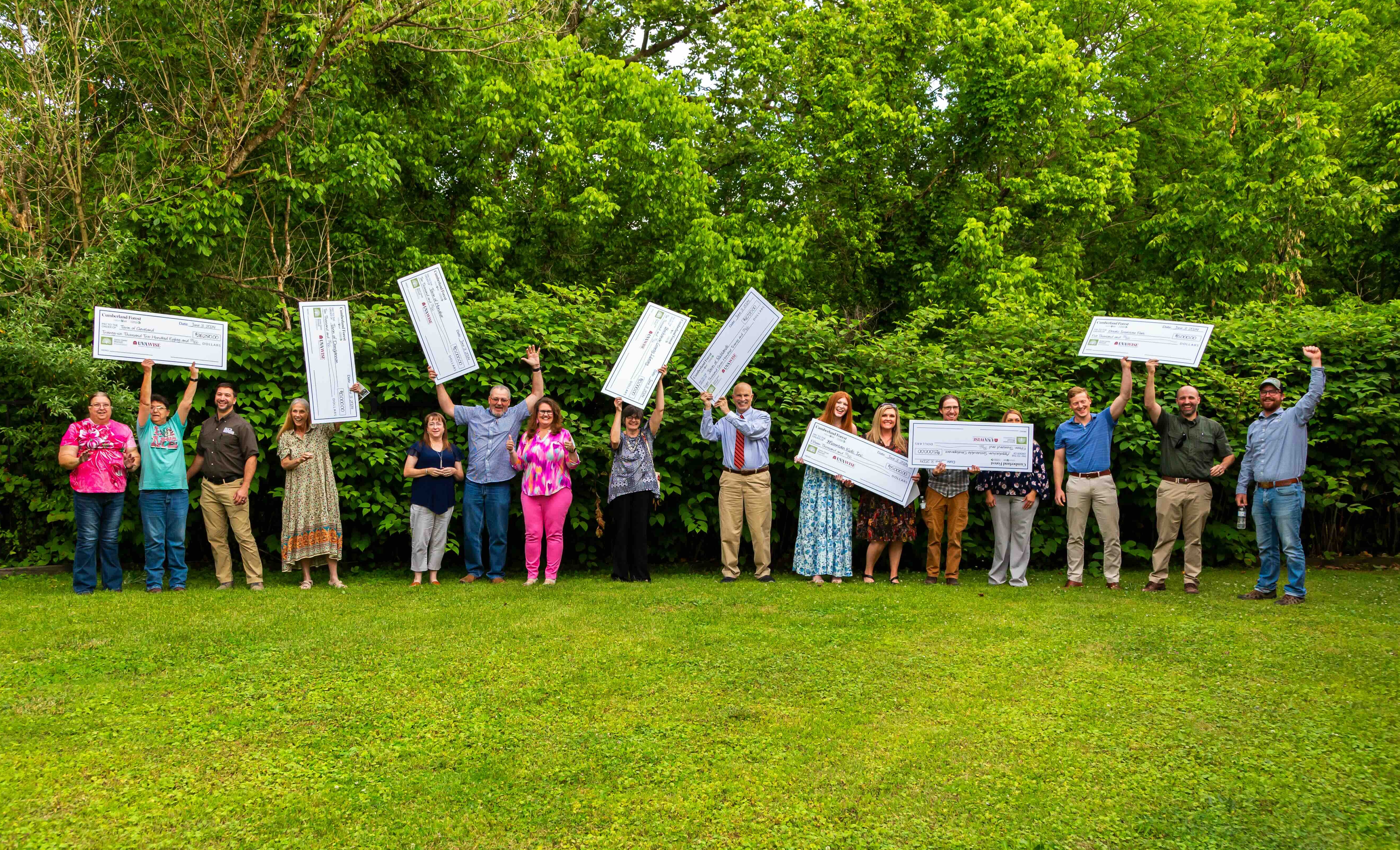 A group of people people pose together outdoors in a grassy era backed by tall trees. Many of the people hold aloft large, novelty checks given out during a grant award ceremony.