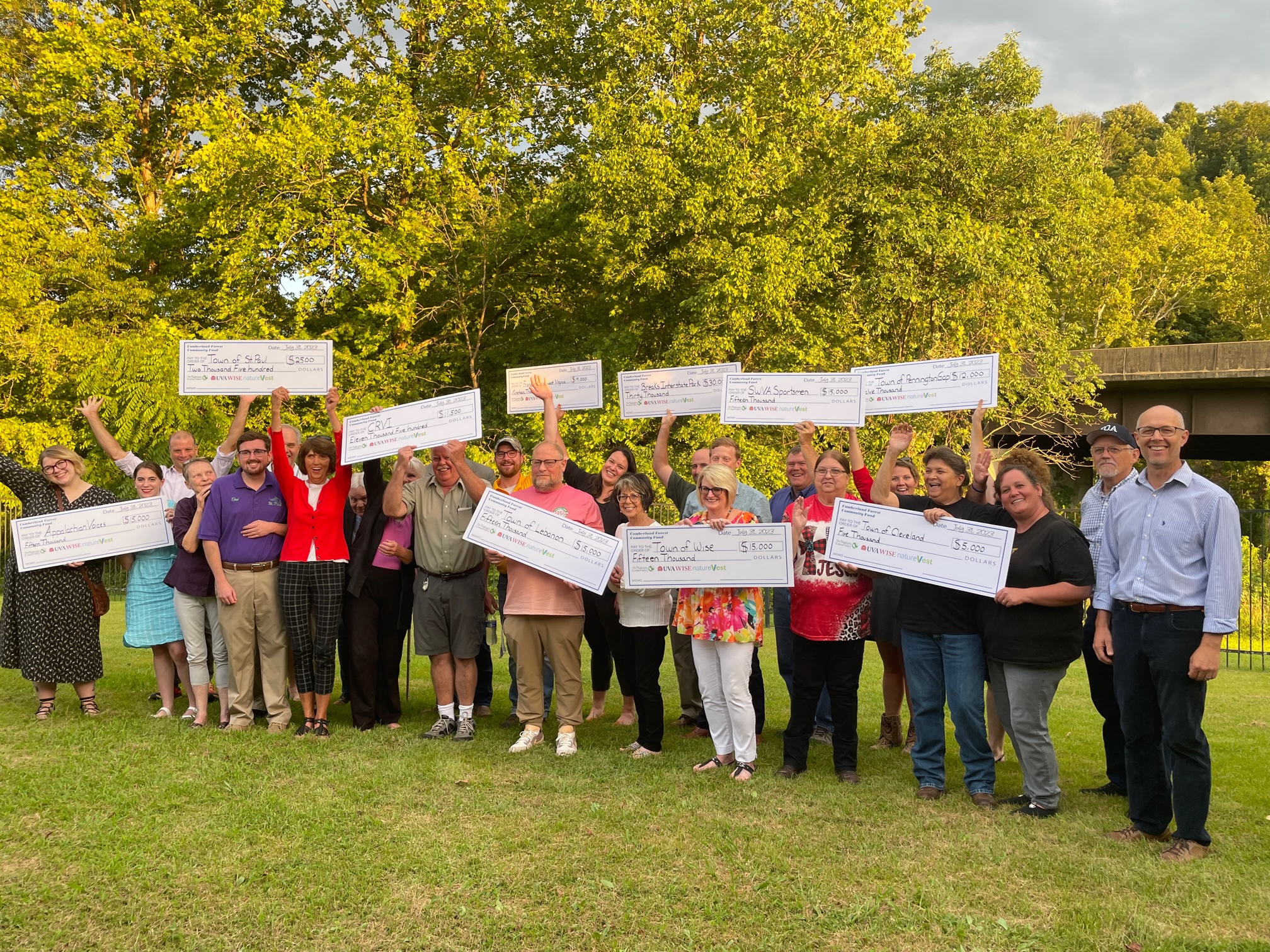 A group of people people pose together outdoors in a grassy era backed by tall trees. Many of the people hold aloft large, novelty checks given out during a grant award ceremony.
