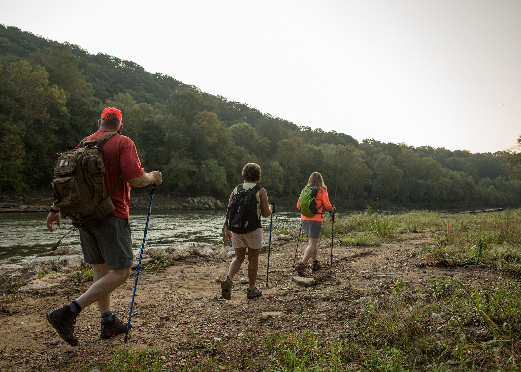 Three people wearing backpacks hike alongside a river.