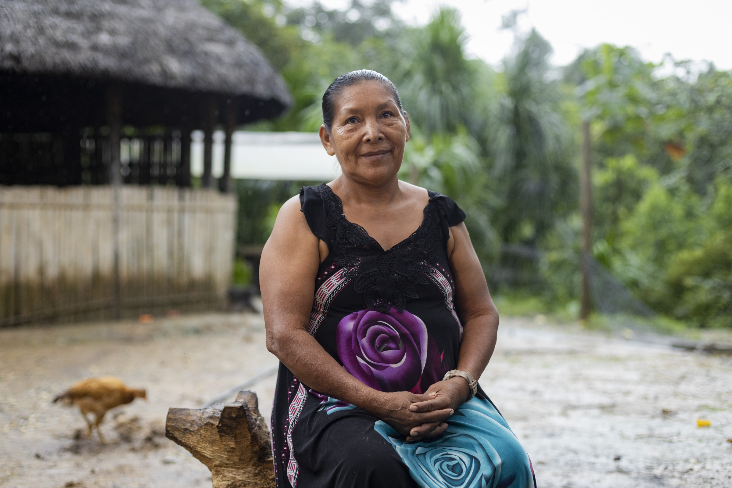 a woman in a black floral dress sits on a stool in her community with a thatched roof building, a rooster and rainforest in the background.