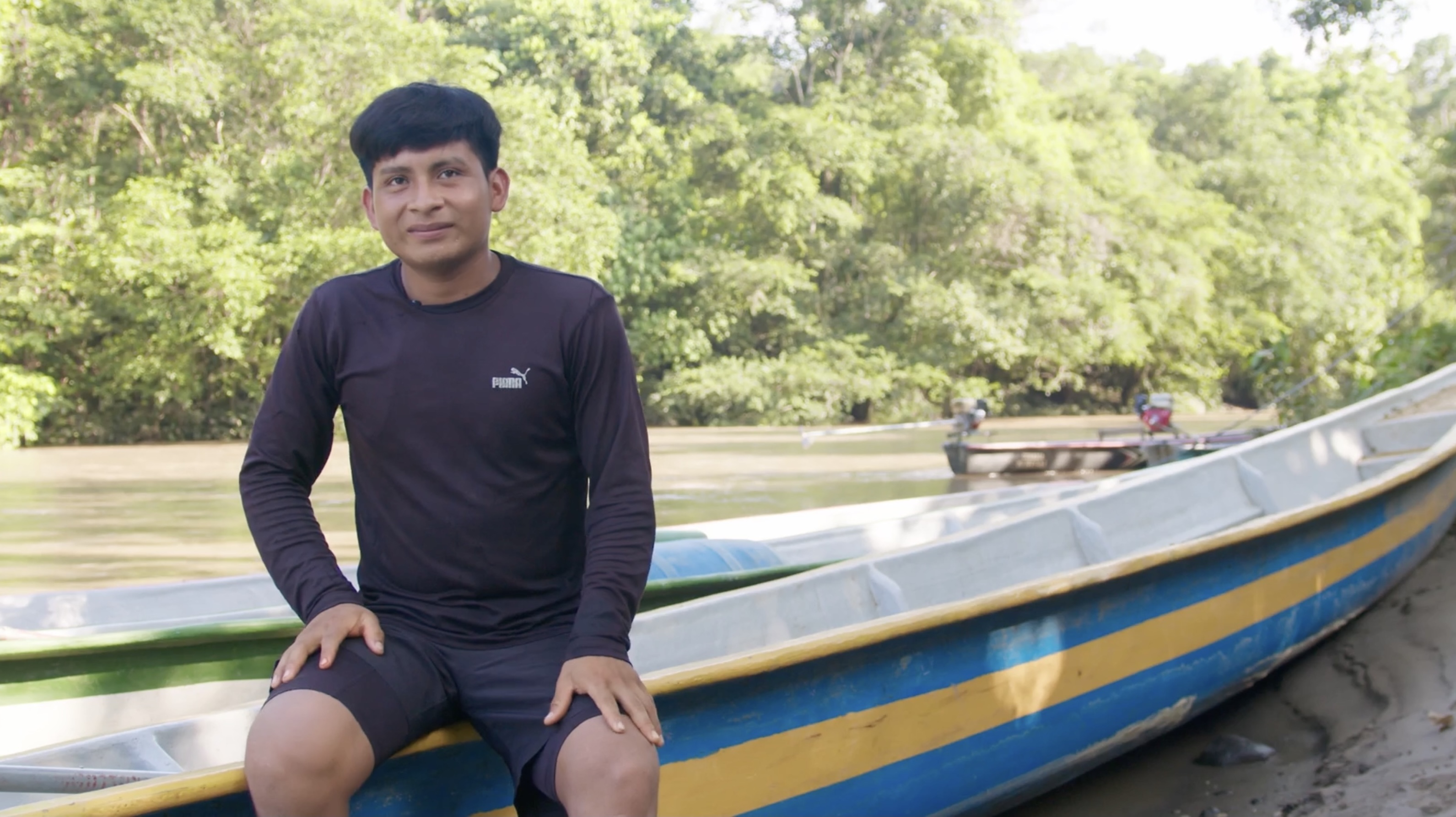 Toribio, a fisherman, sits on the edge of a long narrow boat colored in bold blue and yellow stripes. The boat is on the muddy bank of a river in a rainforest.
