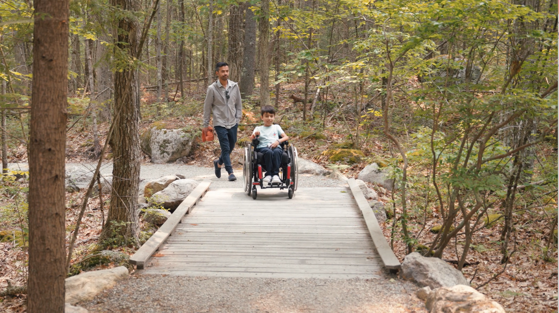 A boy using a wheelchair makes his way down a boardwalk in the woods while his father walks behind him.