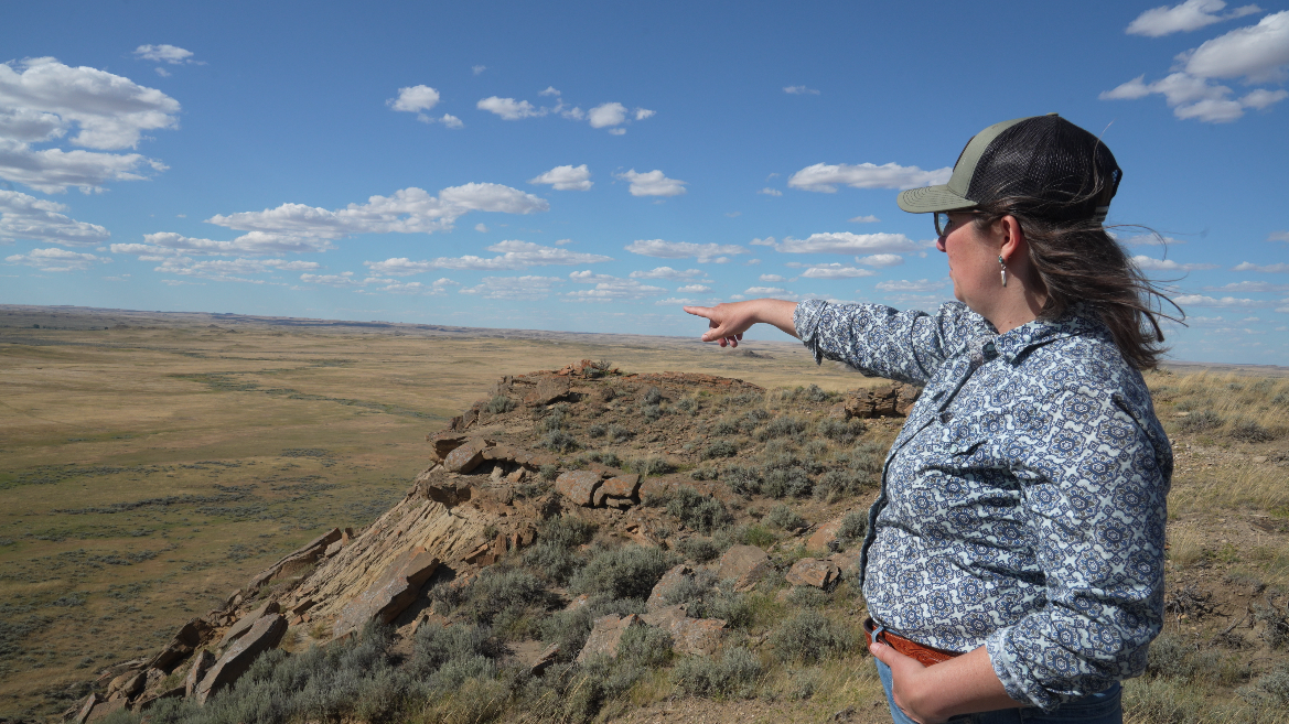 A woman wearing a baseball hat stands on a cliff and points out toward the vast landscape below.
