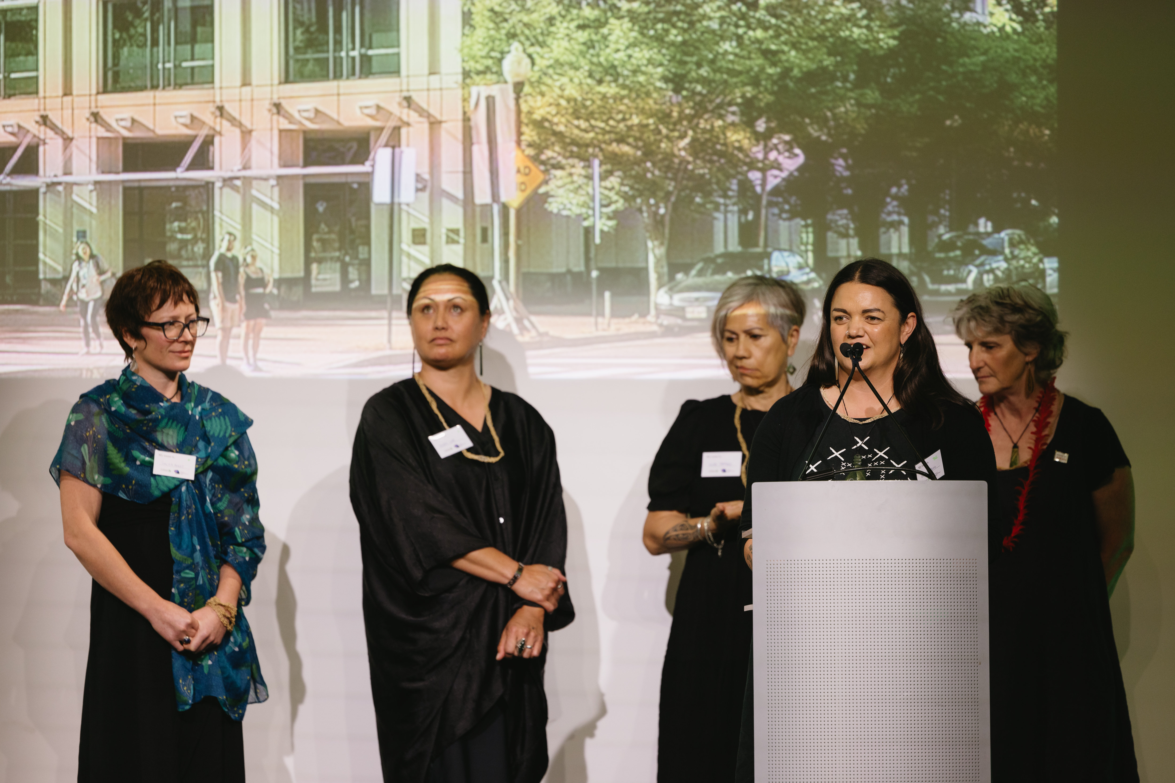 Four woman stand behind another woman speaking at a podium.