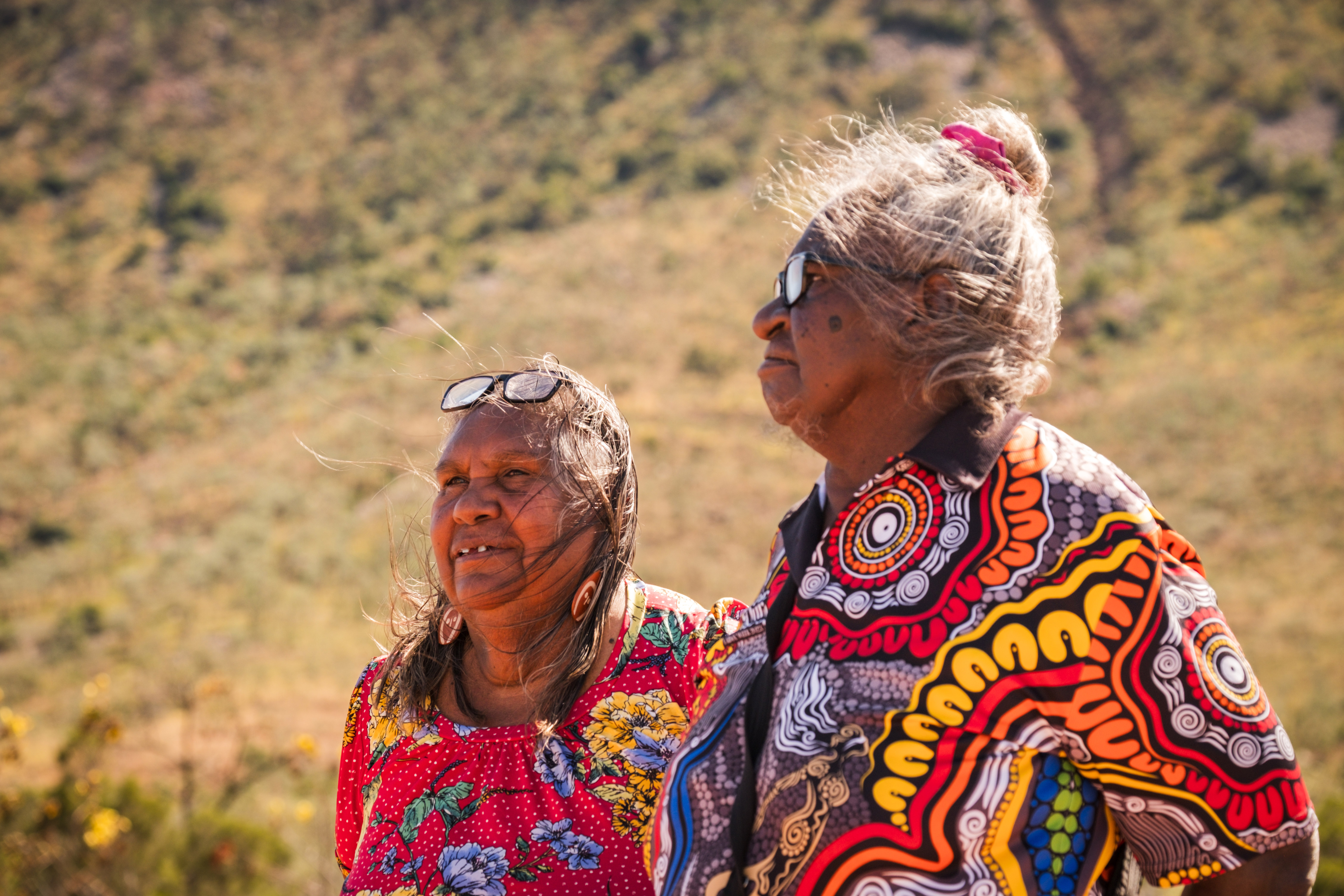 Two women talking to an unpictured third person among the dry scrubby brush of W. Australia.