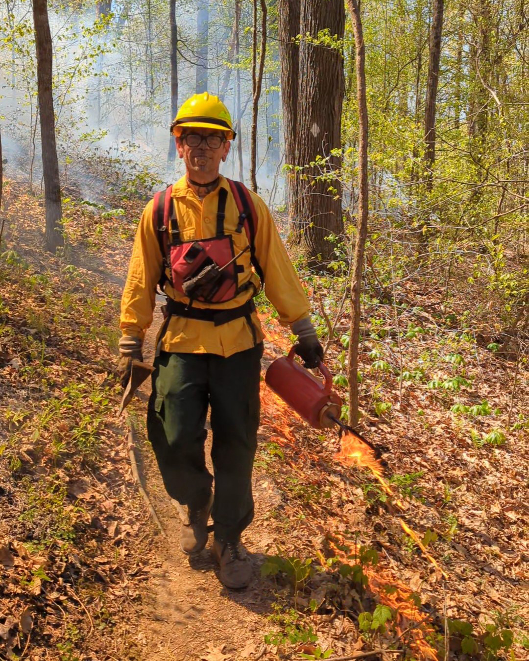 A man wearing yellow clothing and a hard hat walks along a trail carrying a drip torch.