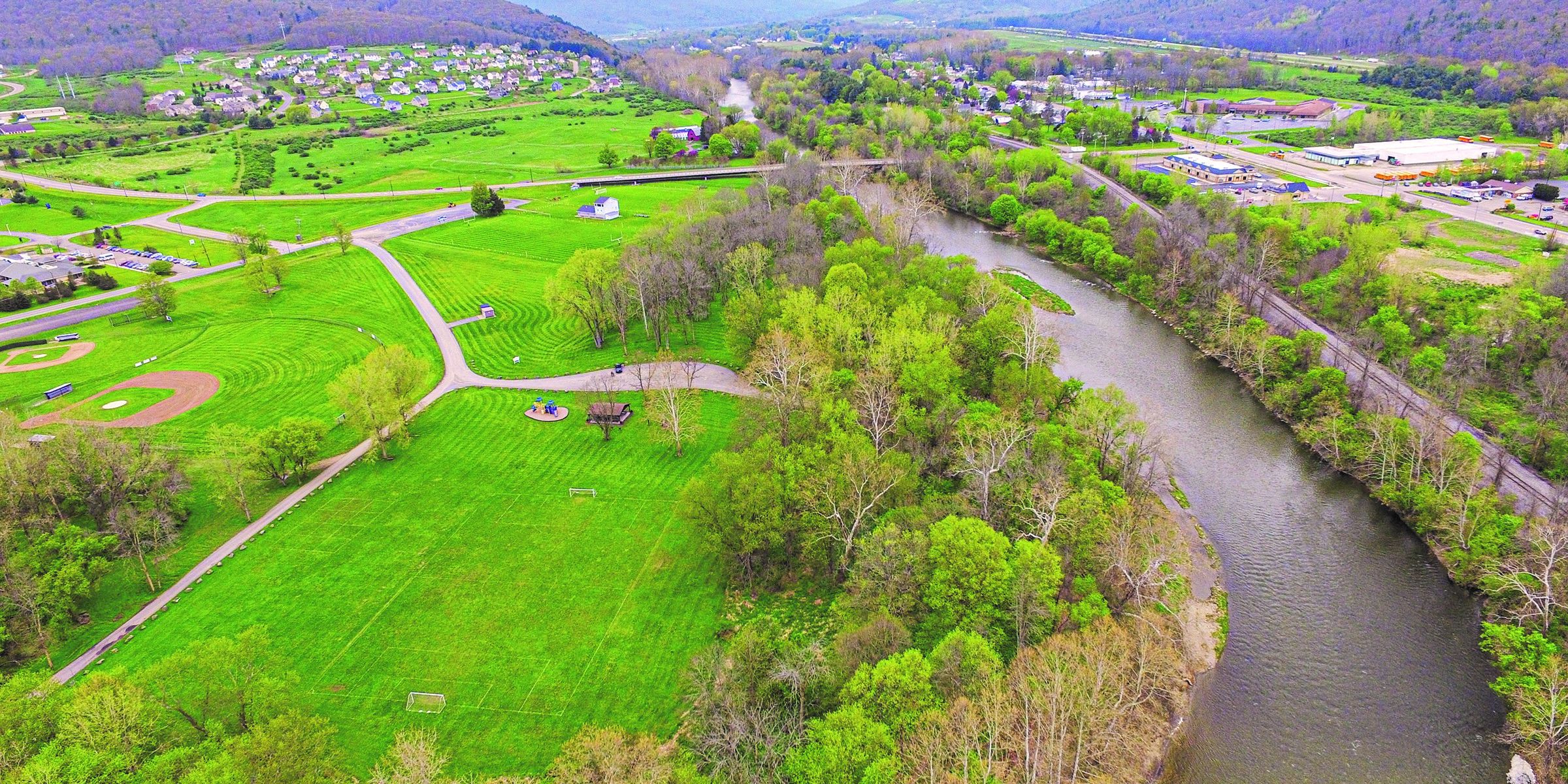  Aerial view of valley studded with green grass and shrubbery on lefthand side, a river cutting down the middle to righthand side, and green and orange shrubbery and homes on the righthand side.