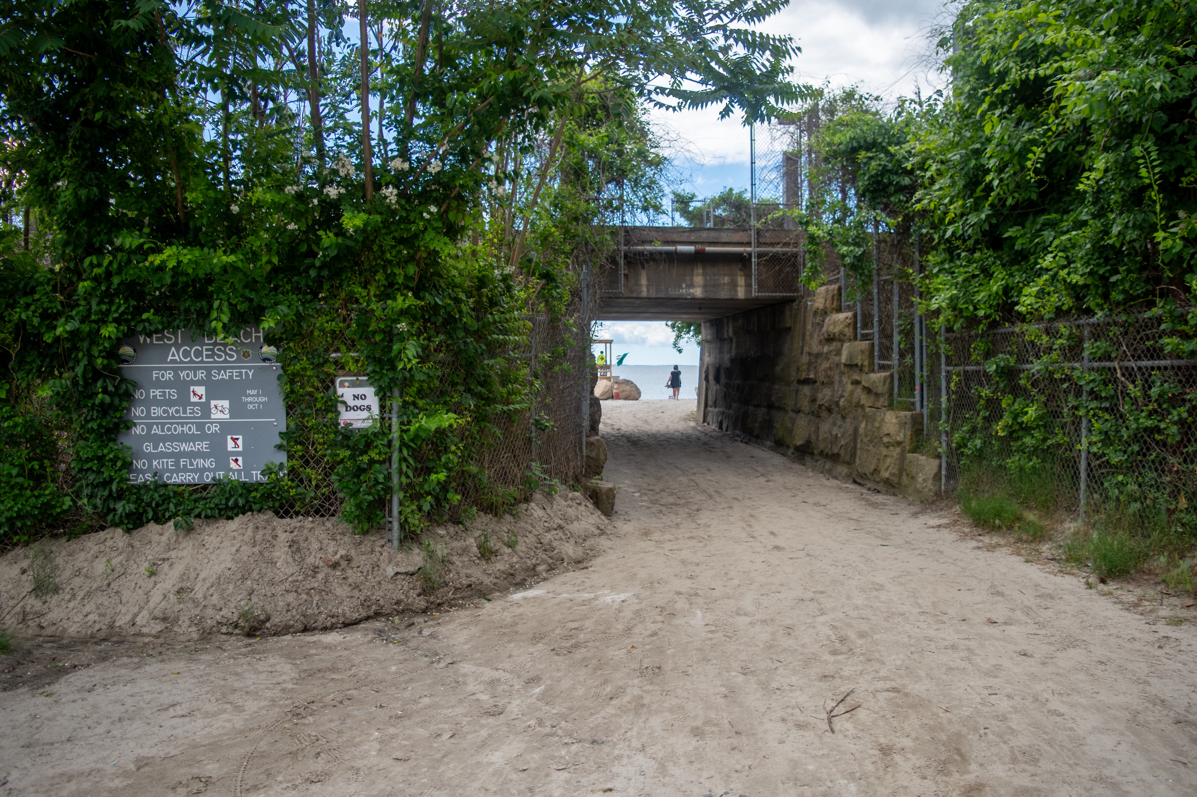 A sandy path leads under a stone bridge toward a beach. The path is lined with lush greenery and a sign listing safety rules. In the distance, a person is seen walking on the beach near the water.