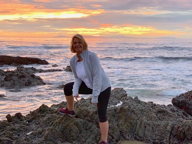 Woman wearing black leggings, white t-shirt and gray jacket stands near an ocean cove with a sunset in the background.