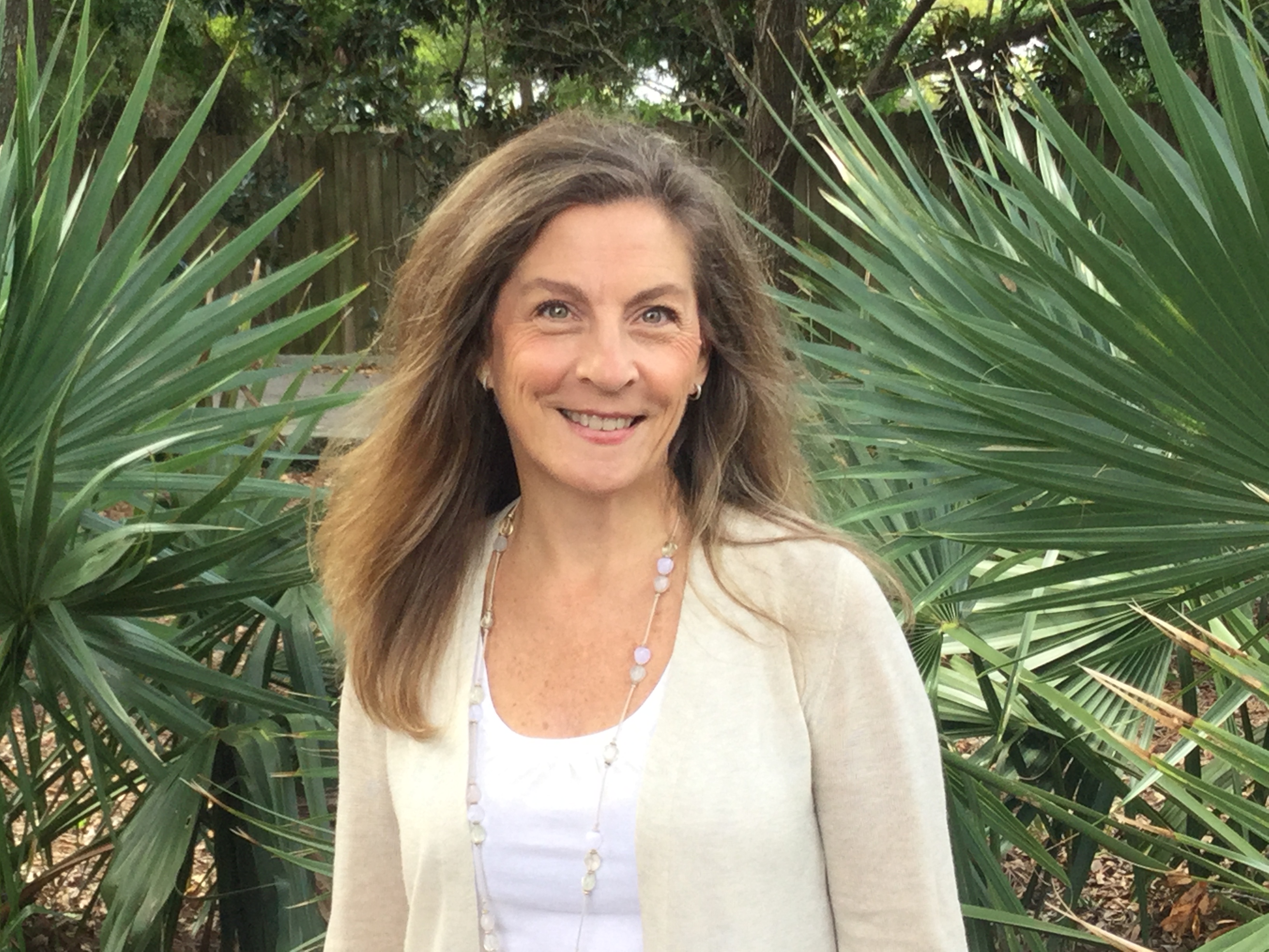 A woman with long brown hair stands in front of large palm tree fronds.