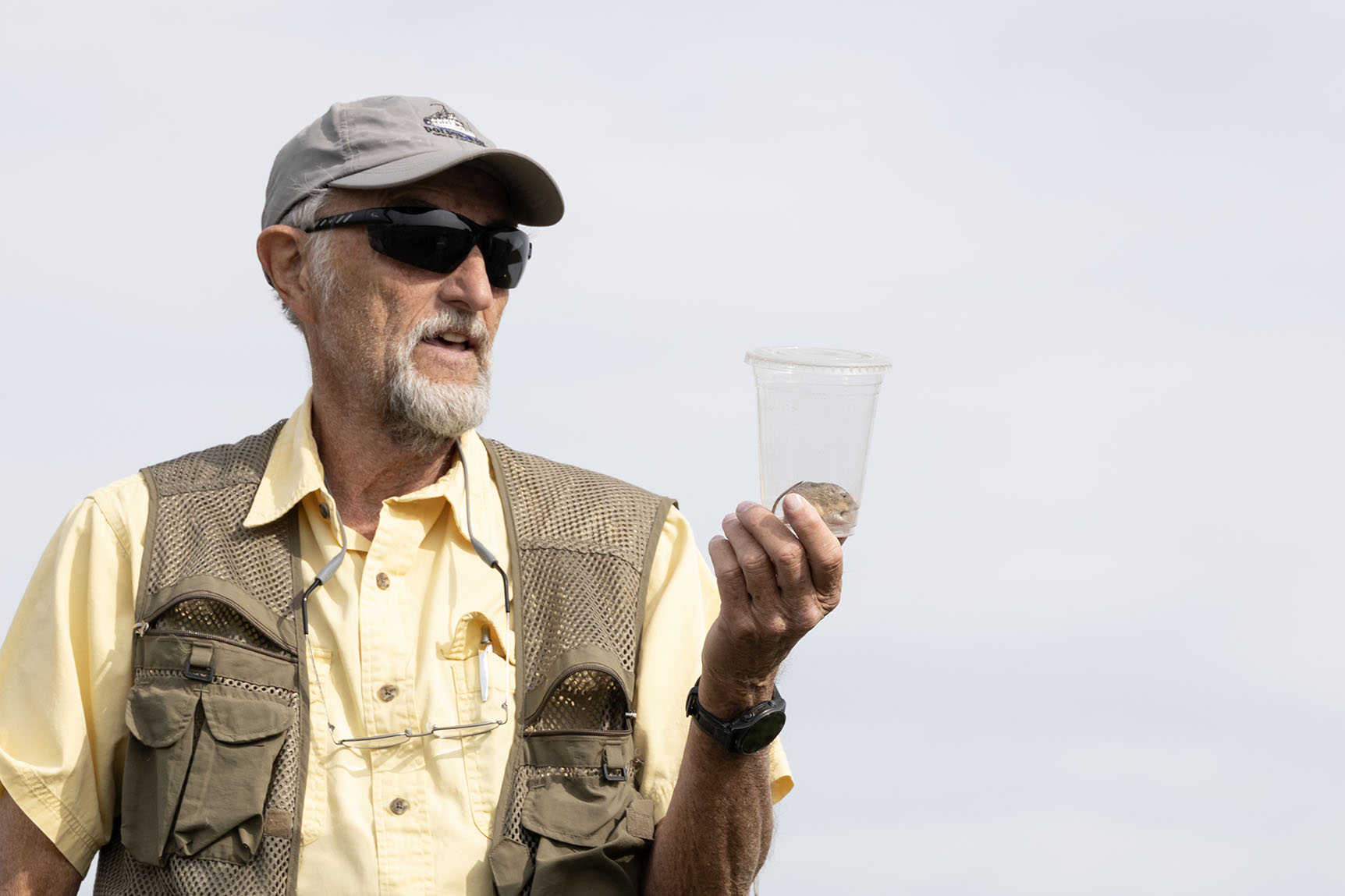 A man in sunglasses and a hat holds up a clear cup containing a small mouse. 