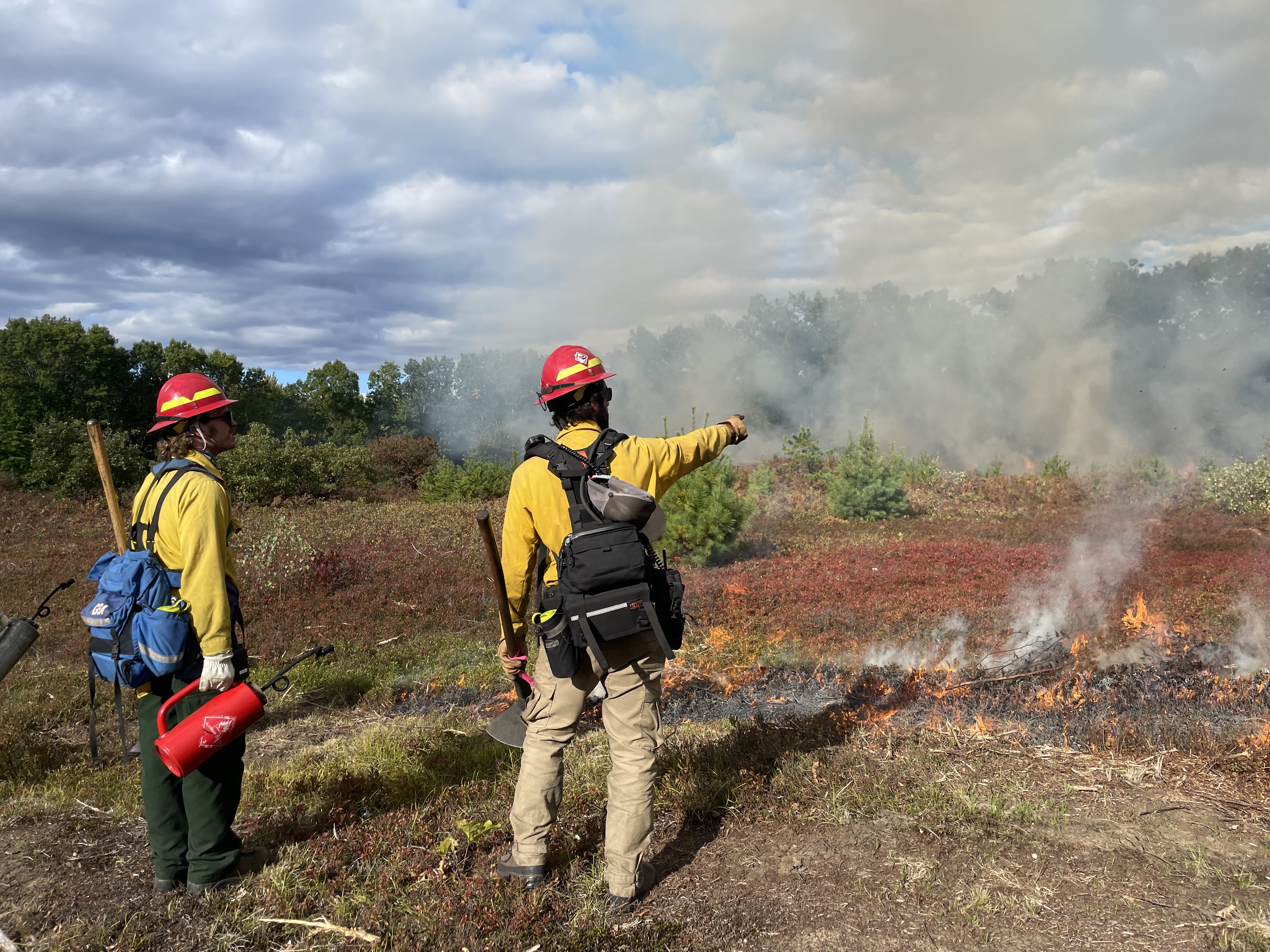 Two people in fire gear stand in front of a small fire in a field pointing into the distance. 