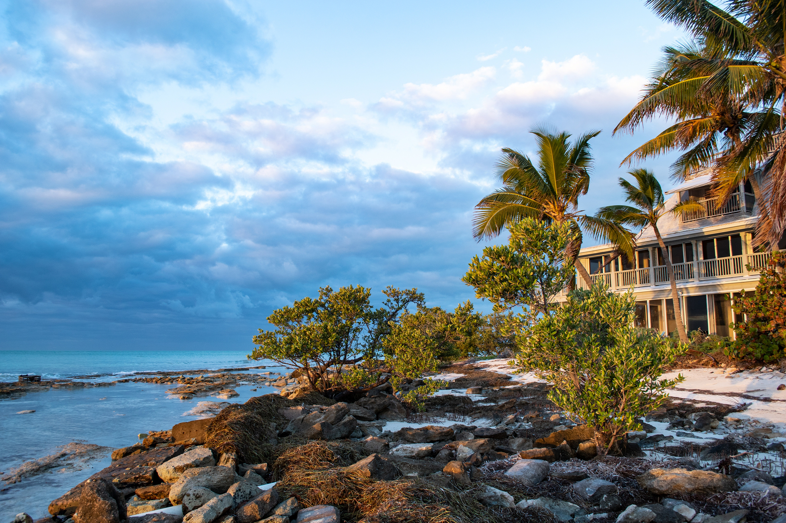 A rocky shore meets the waves; a yellow house in the background.