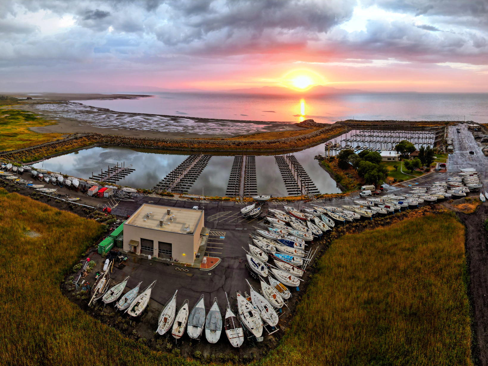 Aerial view of a marina with a lot of boats on dry land, empty docks, and a large expanse of water at sunset.