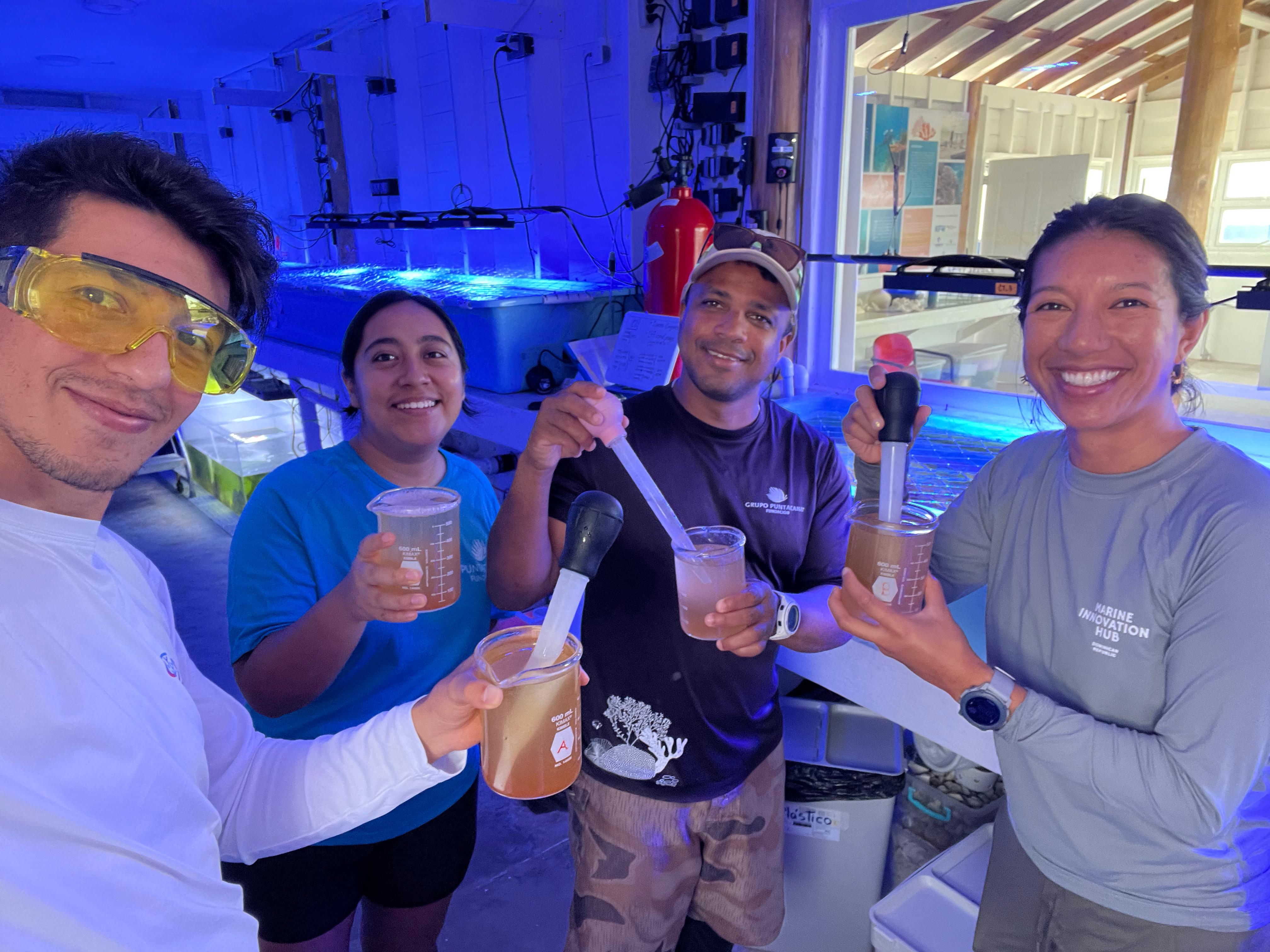 4 people hold lab equipment and smile in a blue-lit room.