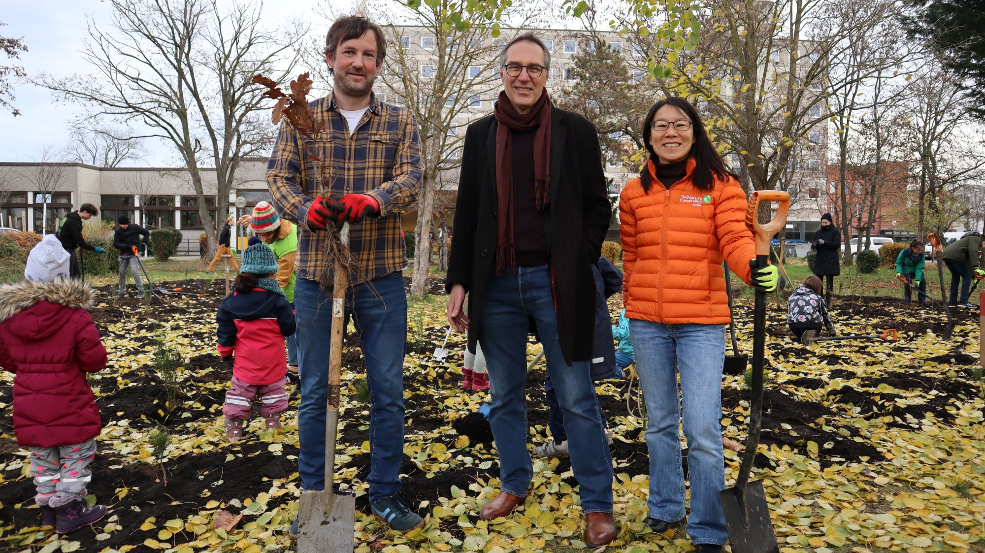 Three people stand in front of a tree planting event with little children running around.