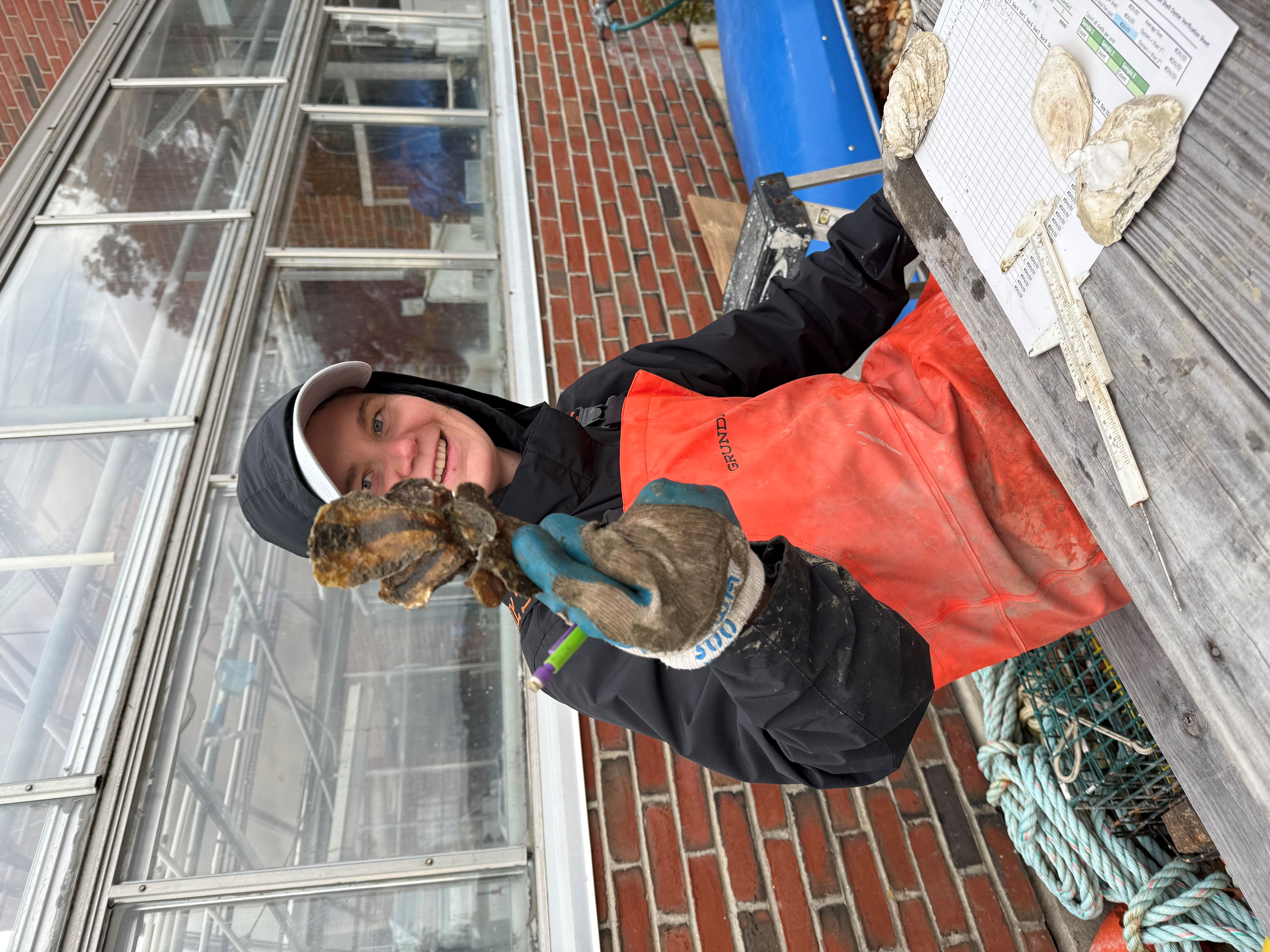 A woman in waders sits at a picnic table and holds a clump of oysters.