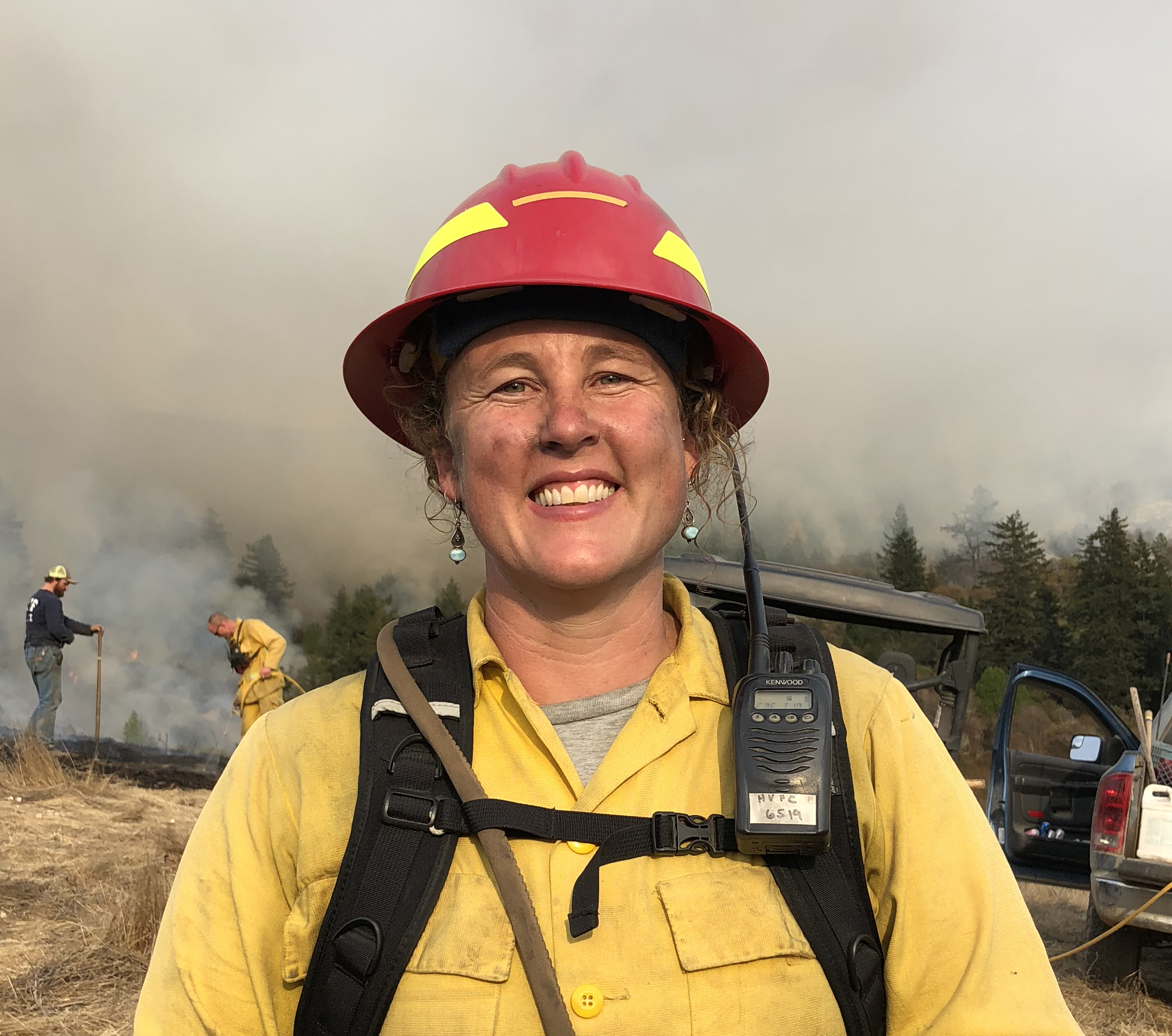 Women in yellow fire gear & red hardhat stands by prescribed burn.