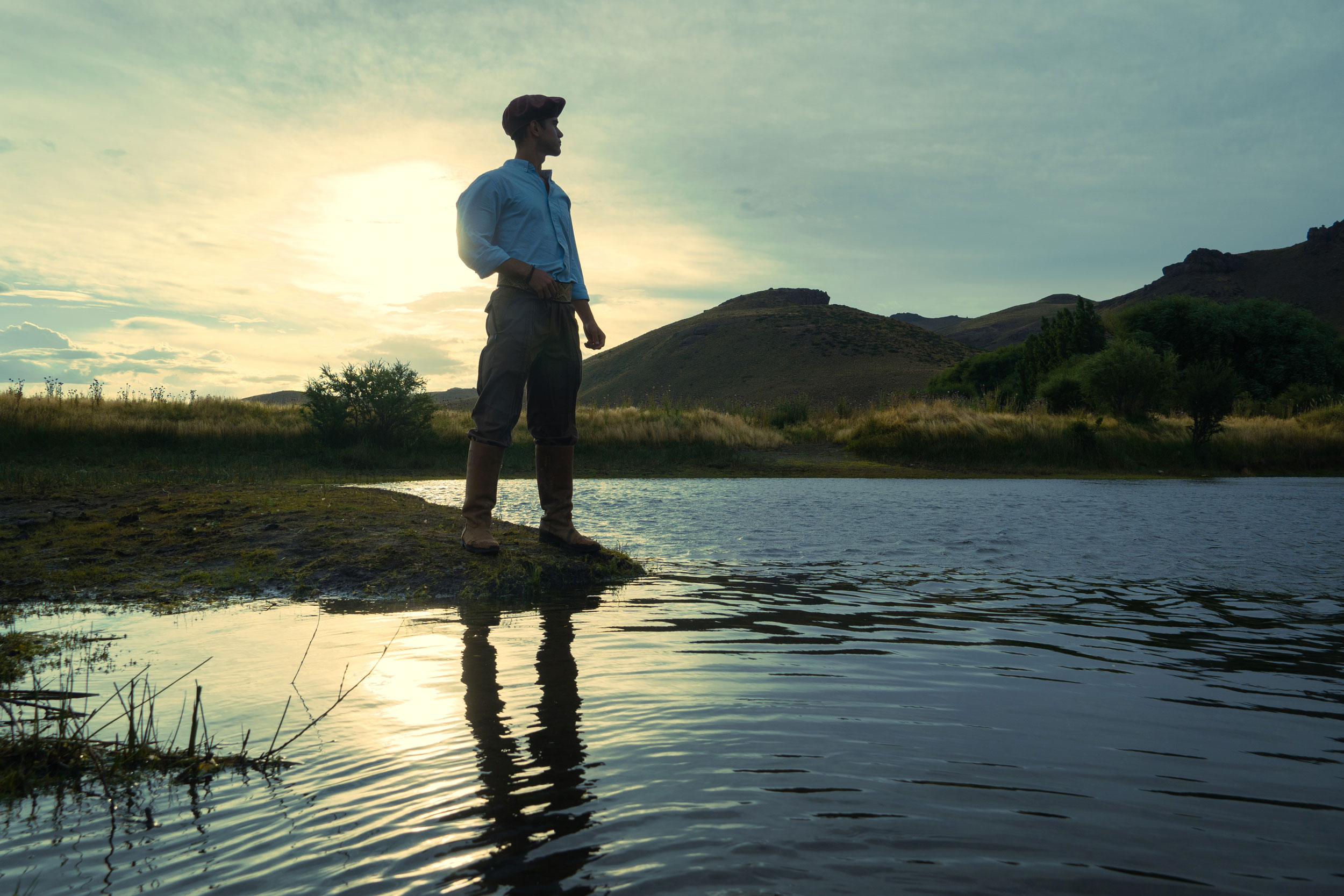 Un hombre posa en una roca cerca de un lago tranquilo, con árboles y montañas visibles en el fondo.