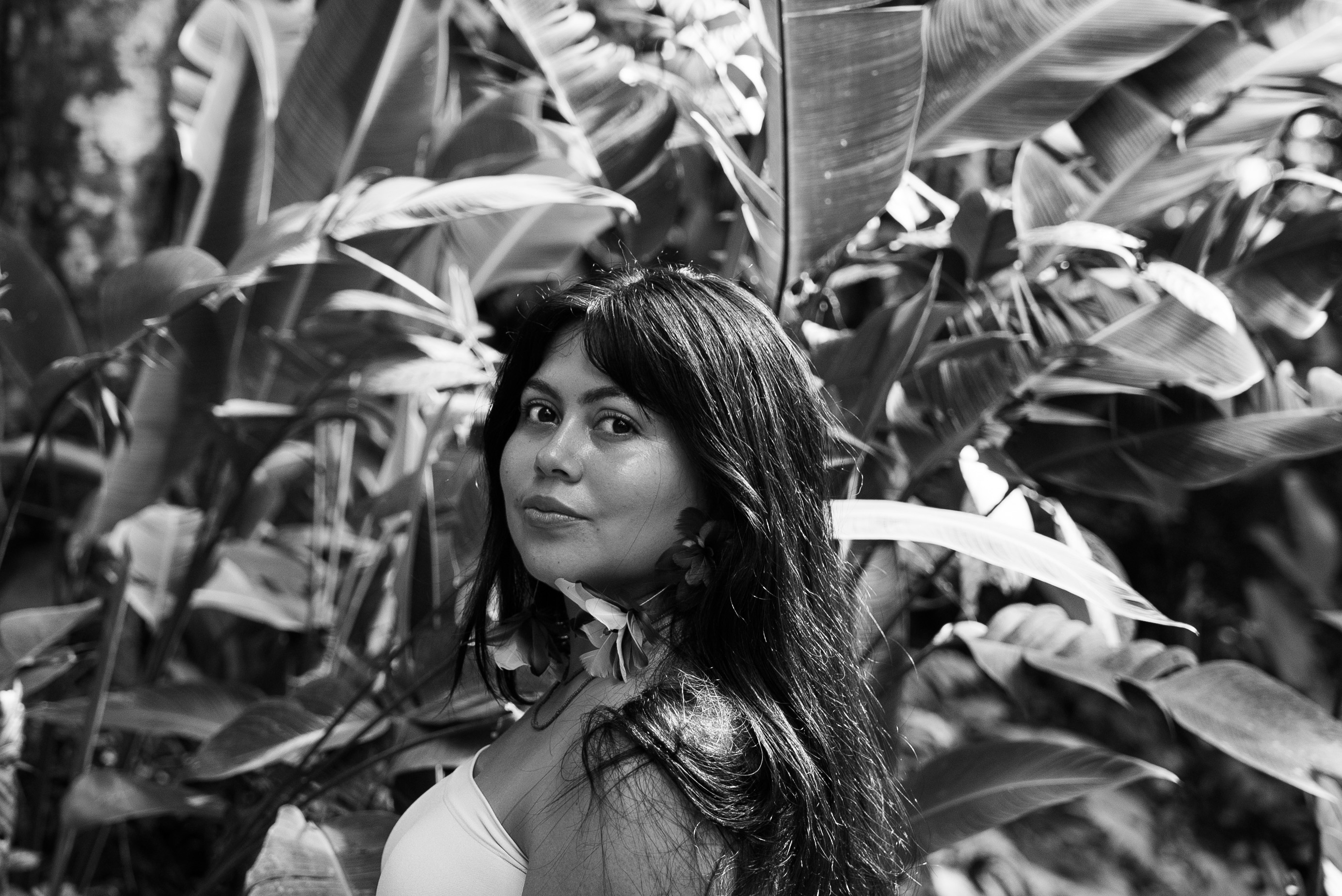 Retrato en blanco y negro de Eli Virkina, fotógrafa, observando a la cámara de perfil, con un fondo de selva. 