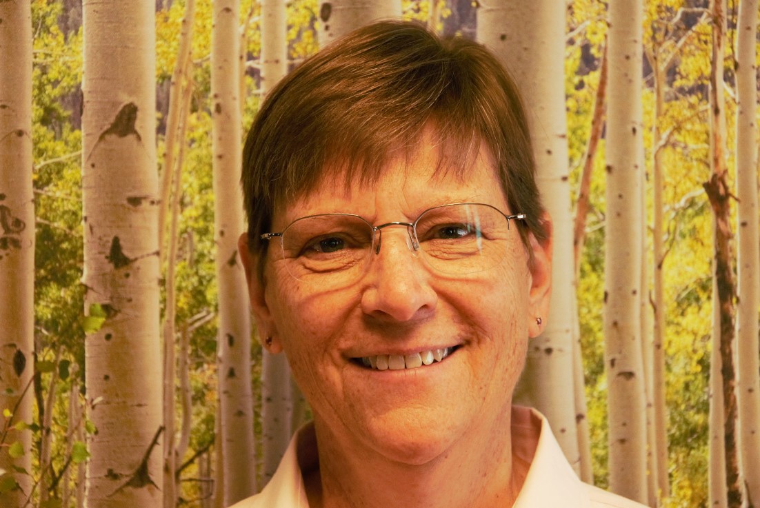 Woman in glasses smiles in front of aspen tree backdrop.