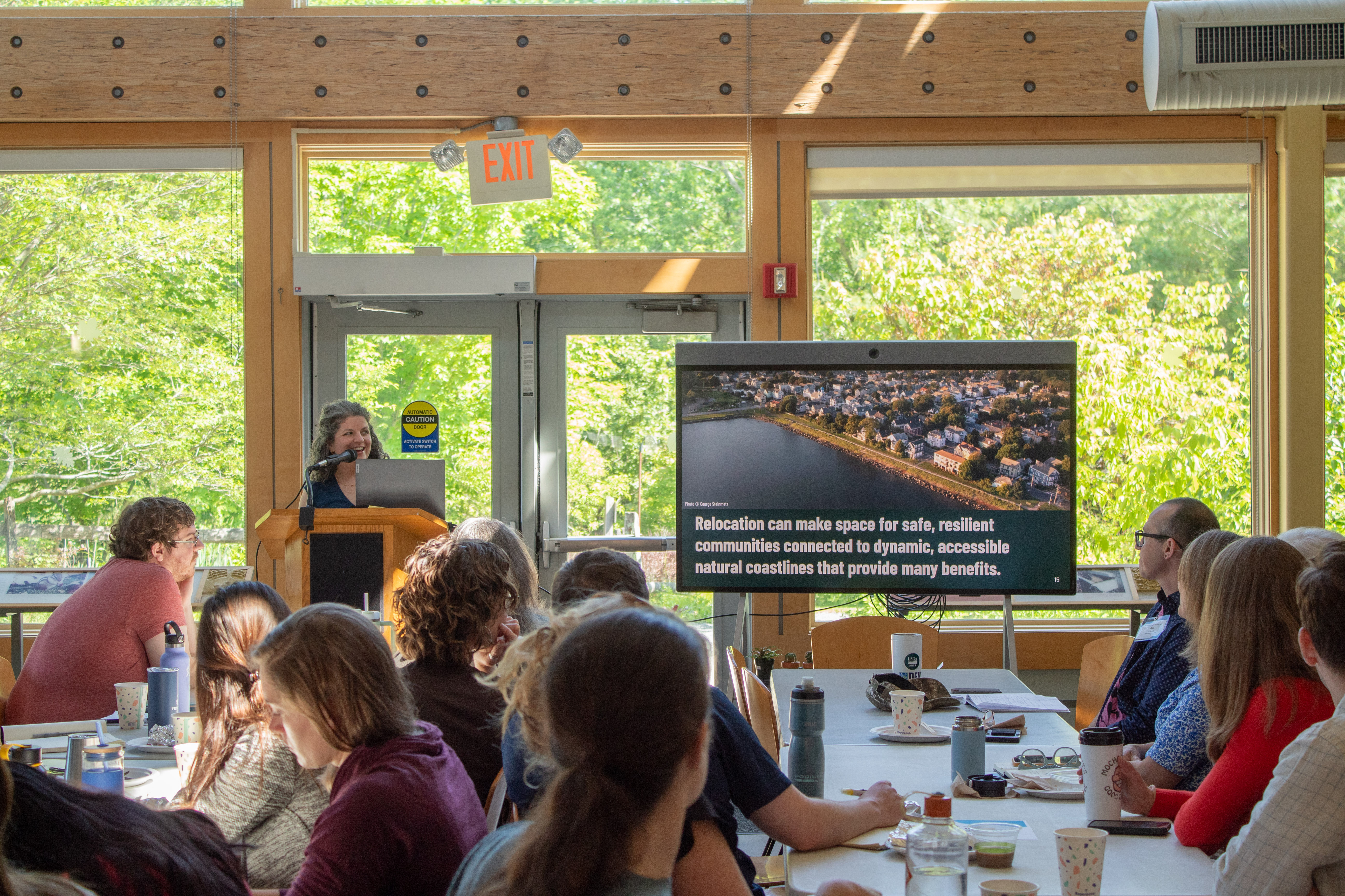 A TNC staff member speaks at a podium with a group of people facing her out of focus. There are slides on the screen at the front of the room. 