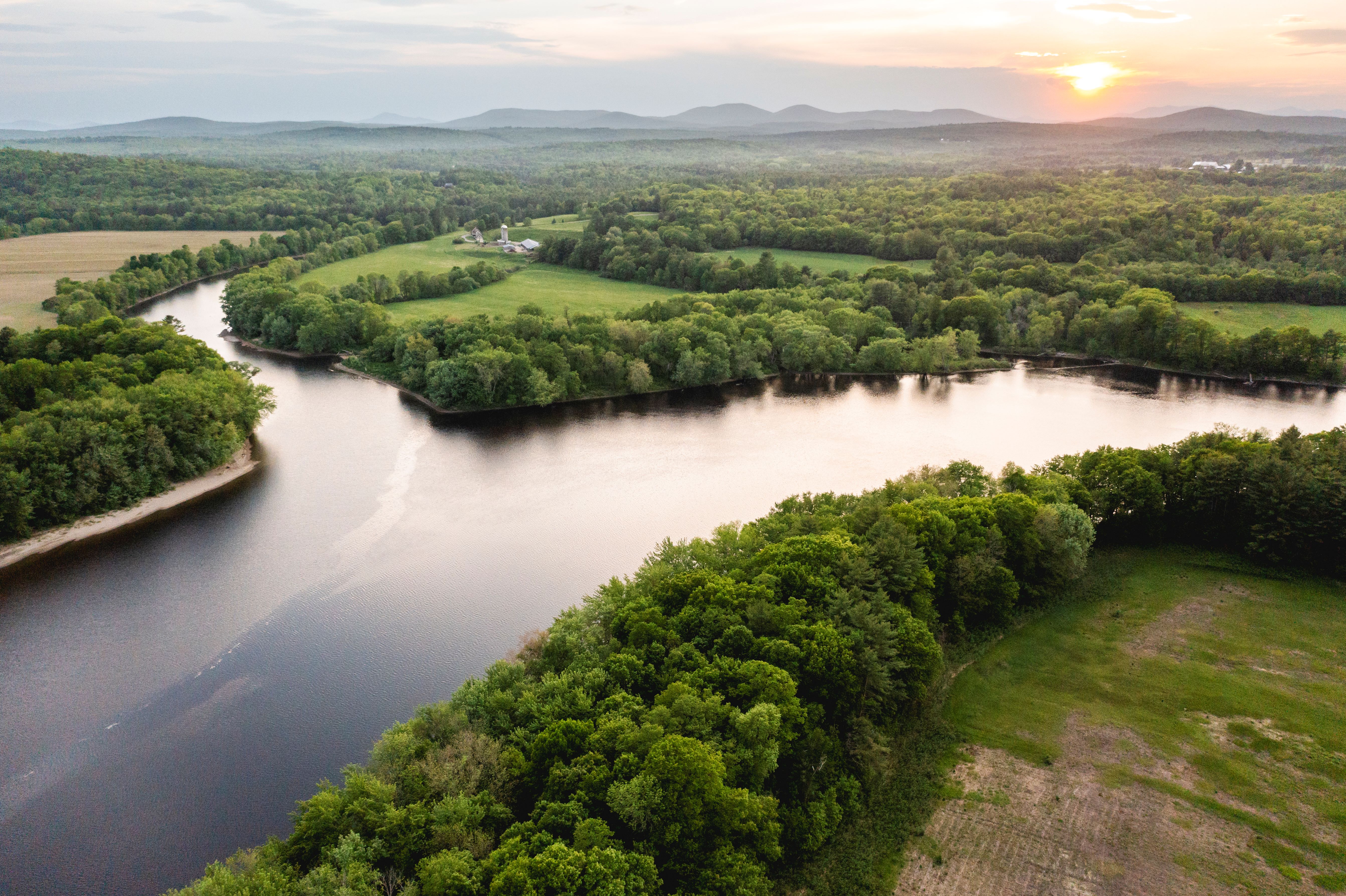 A wide river splits into two branches, flowing past tree lined banks and open fields.