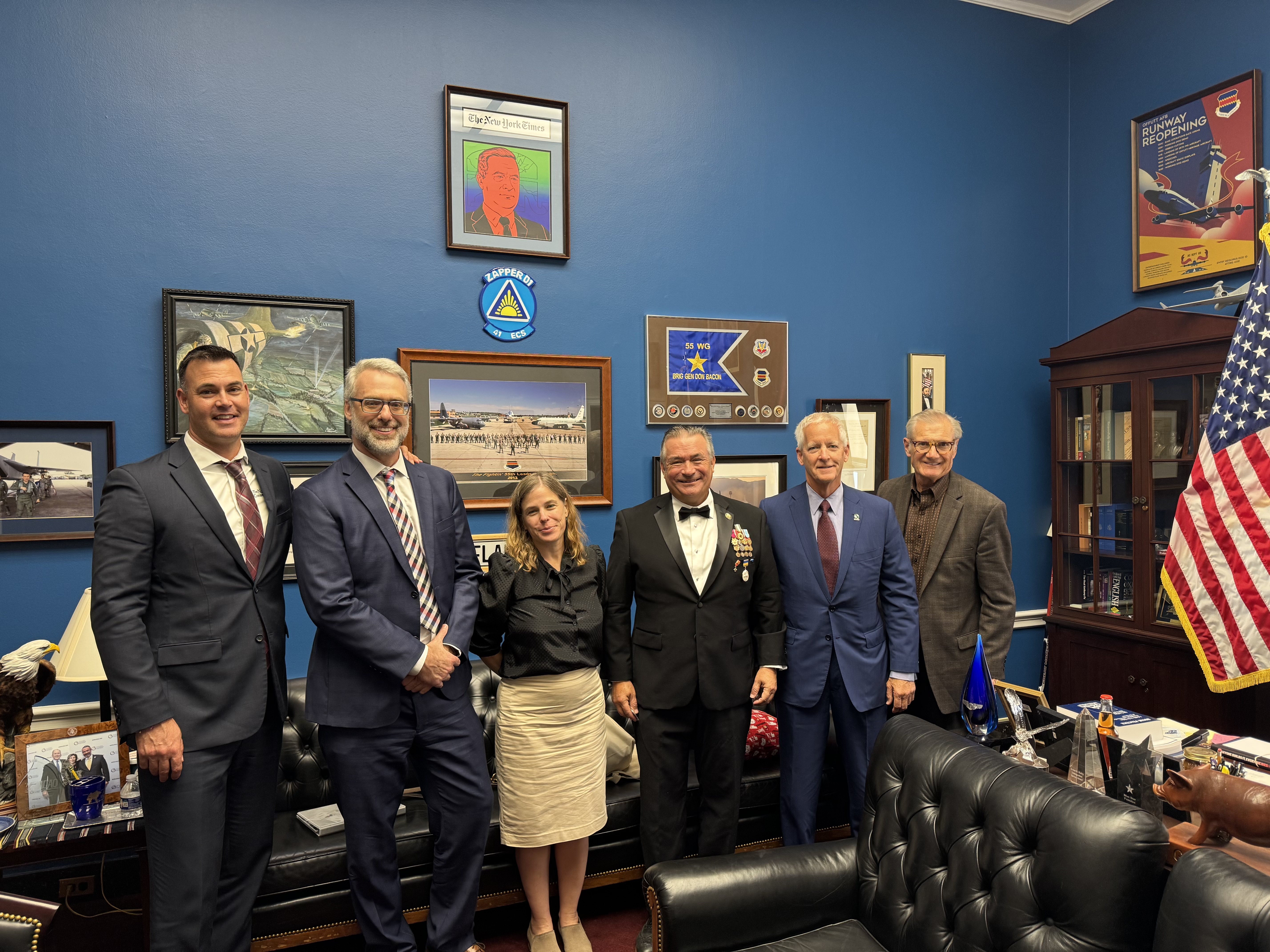 A group of people pose in front of a wall of images and plaques in a Congressman's office.