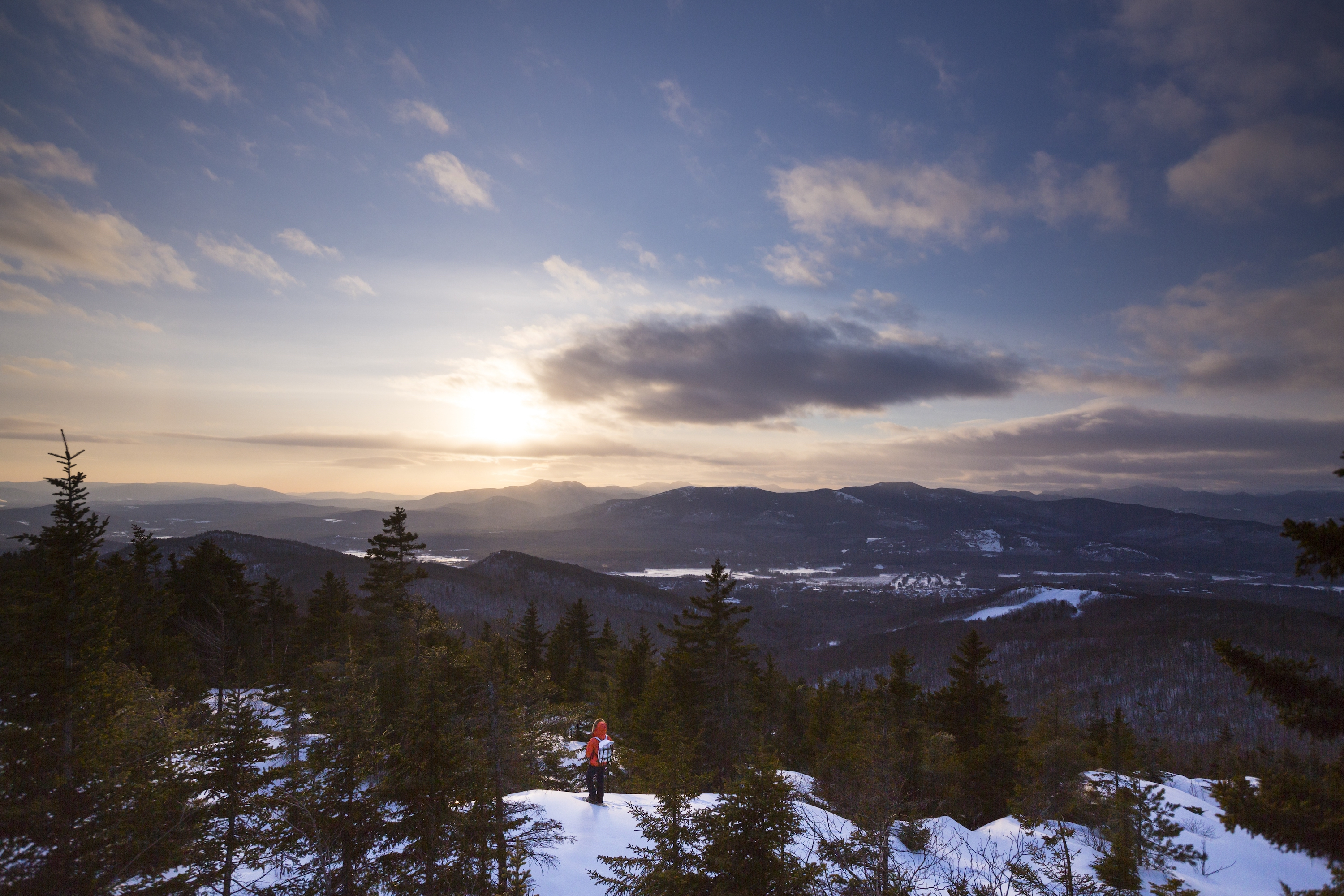 A woman in a red winter coat and white backpack stands on a snowy ridge and looks out over a mountainous vista.