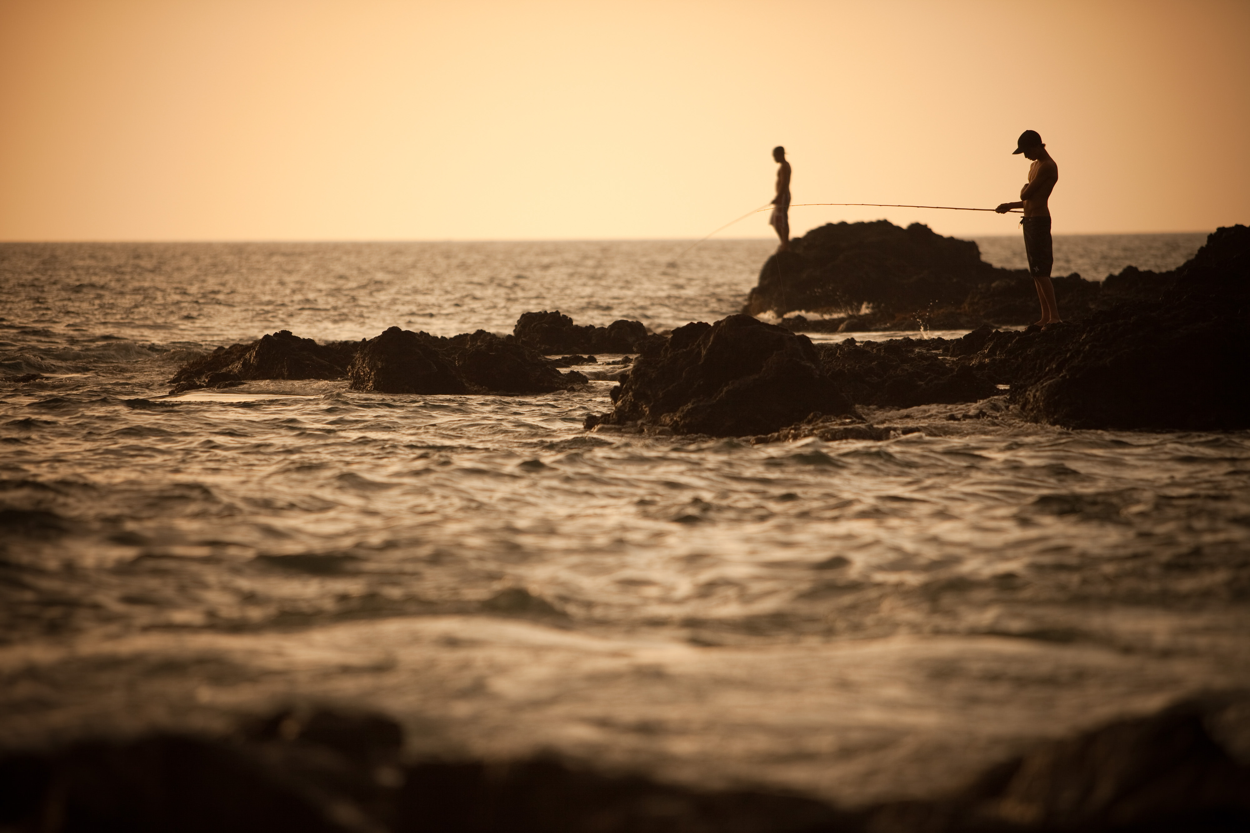 Two fishermen stand on a rock surrounded by ocean.