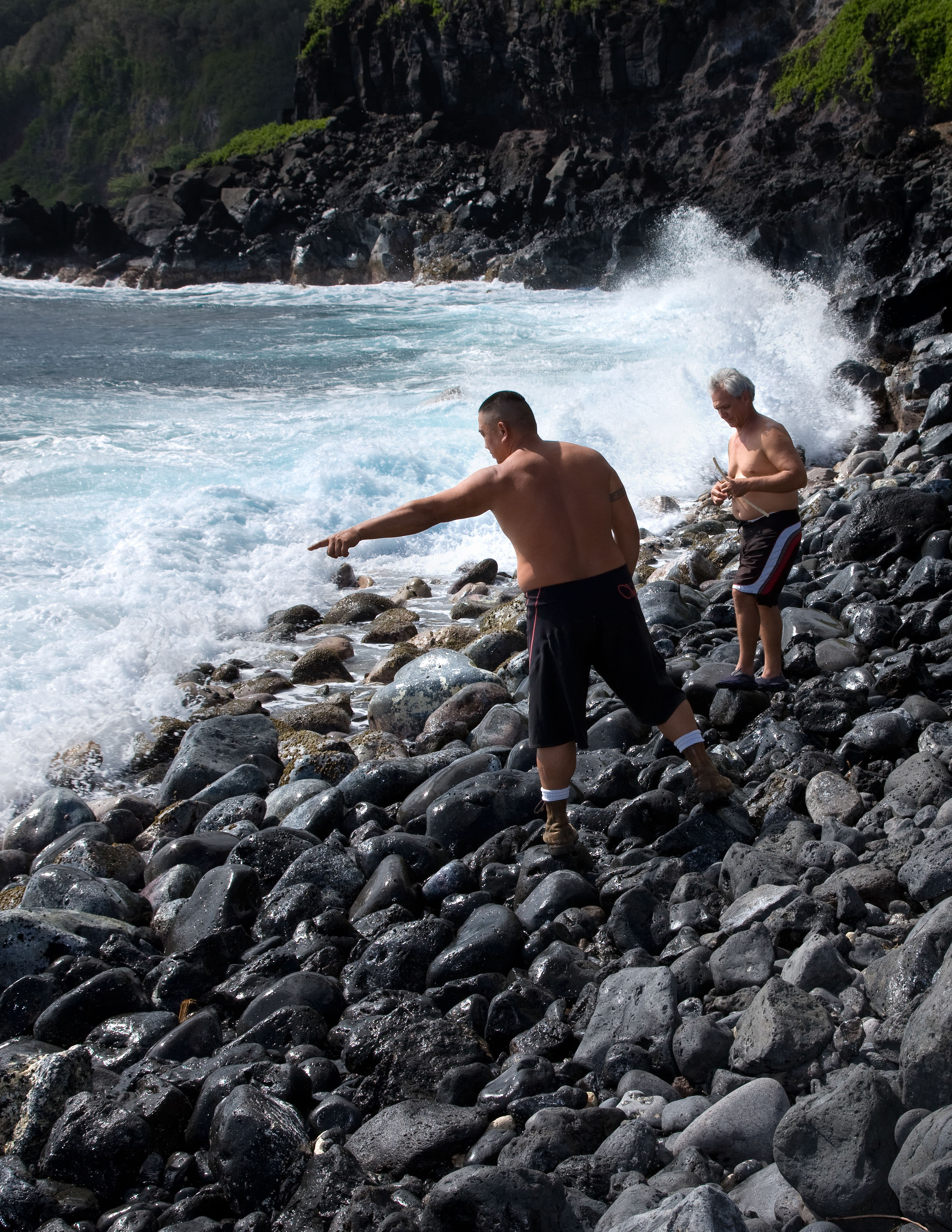 Two men stand on a rocky beach near crashing waves.