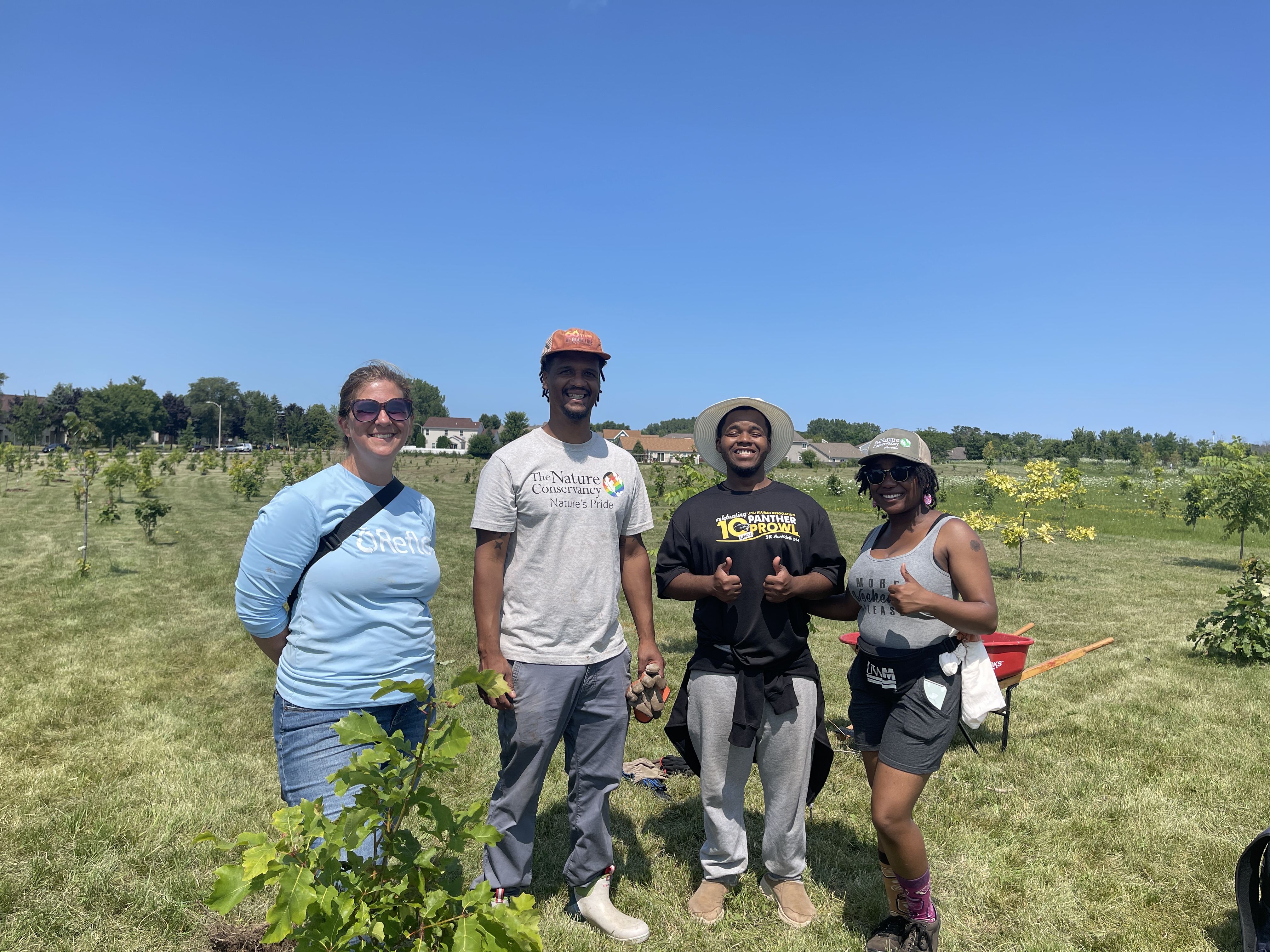 Four people stand outside in a large green space with small, newly planted trees around them.