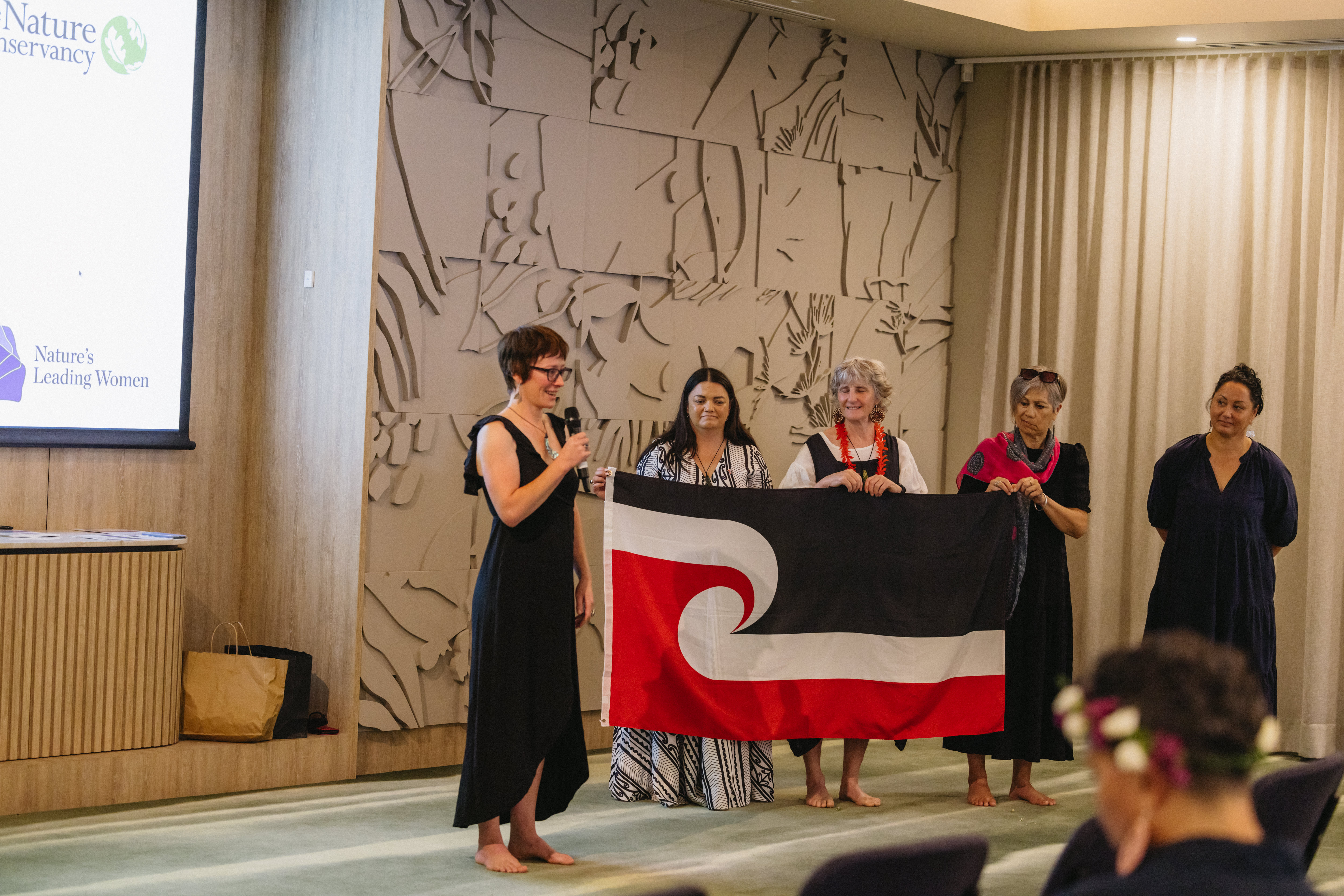 Four women hold a flag while another woman speaks into a microphone.