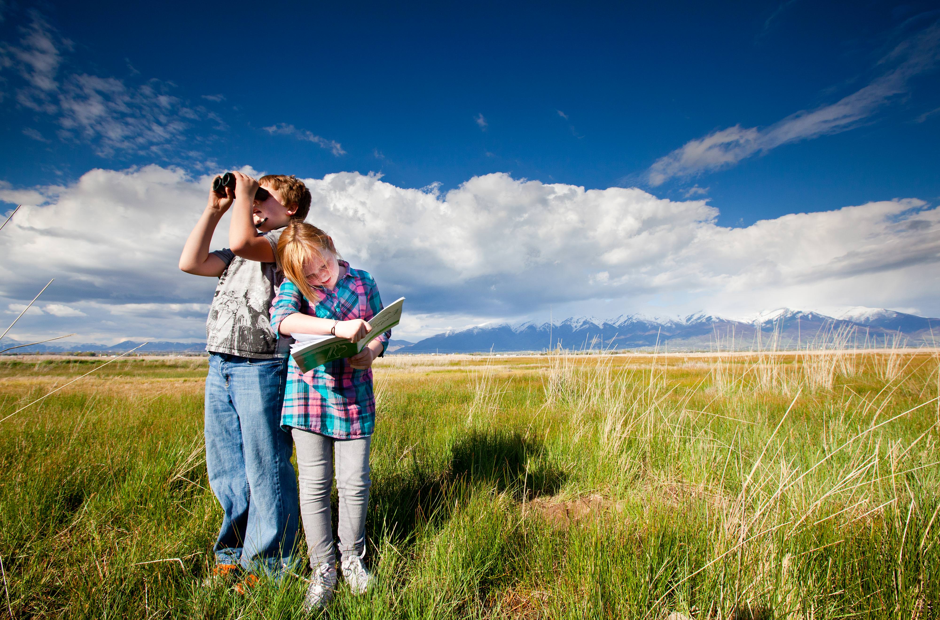 Two kids standing on grass and looking through binoculars.