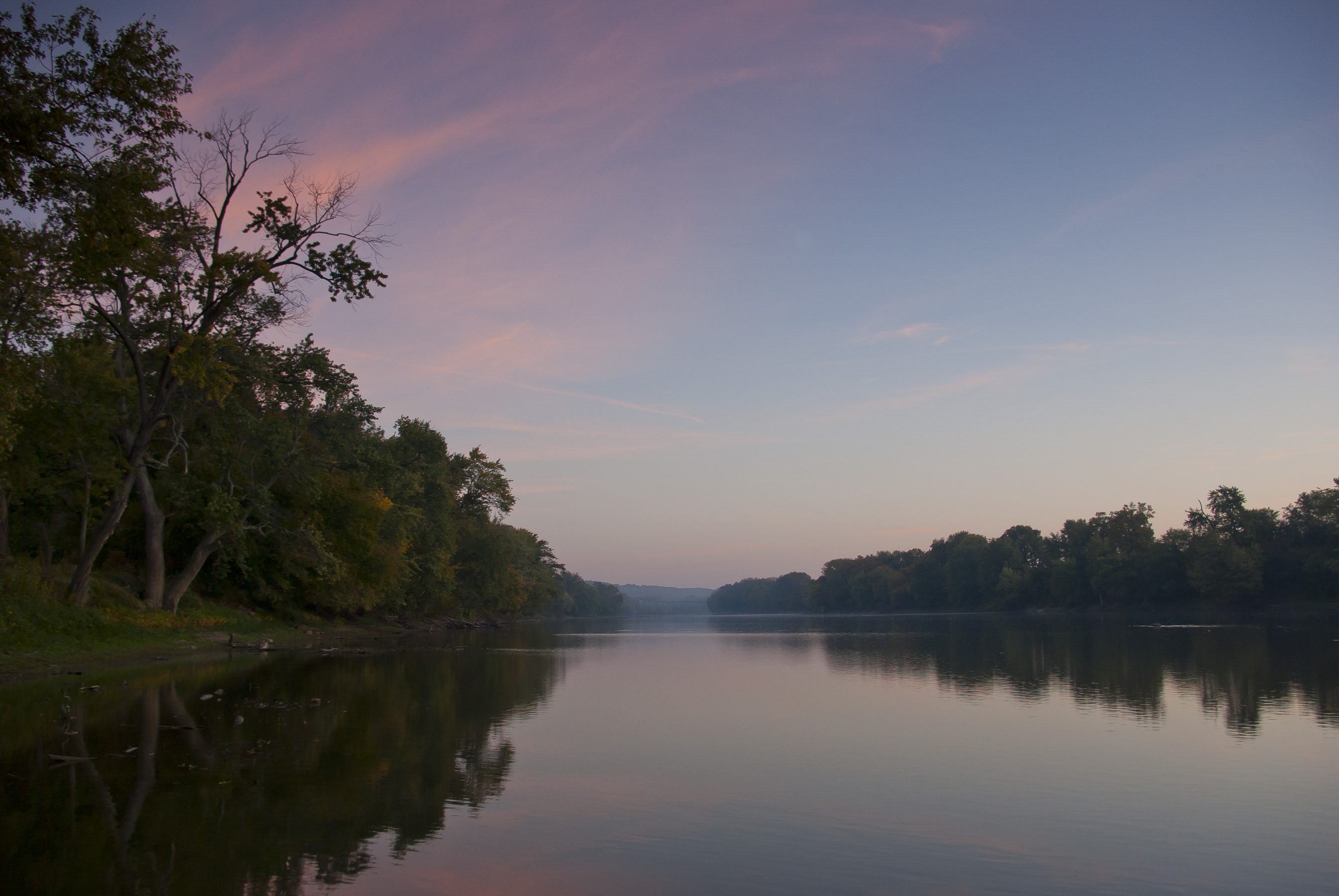 A view down a calm river with dusky pink sky and mist over the water.