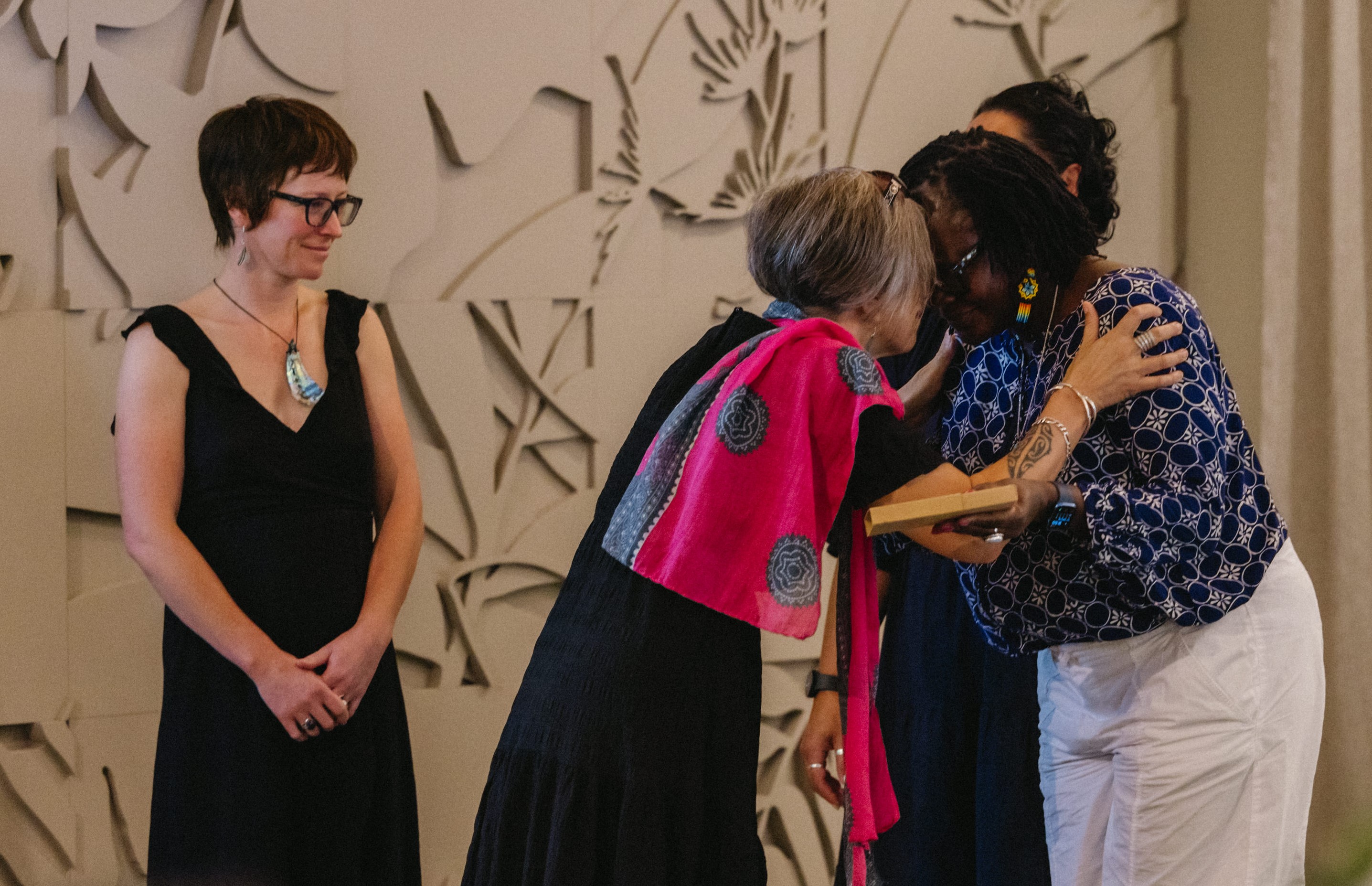 A woman wearing a colorful scarf embraces another woman on a stage. 